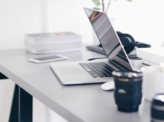 A half-closed laptop on a busy desk with headphones, camera lenses and books