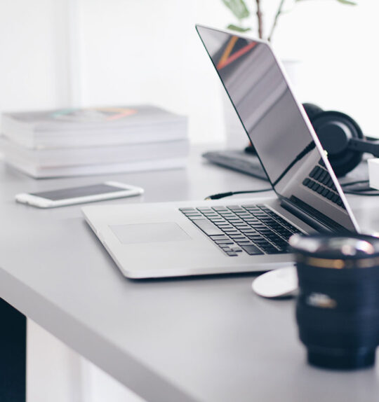 A half-closed laptop on a busy desk with headphones, camera lenses and books