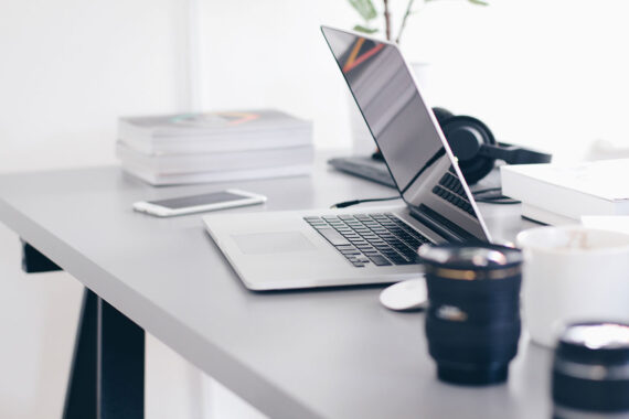 A half-closed laptop on a busy desk with headphones, camera lenses and books