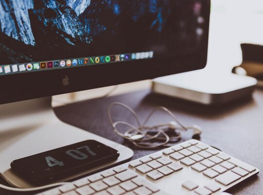 Close-up of a desk with an iMac and a smartphone displaying the time